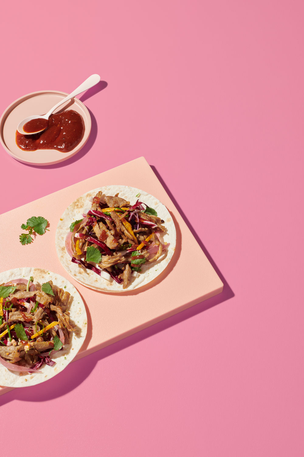 Two plates of planted.pulled tacos on a pink background, with a small dish of sauce